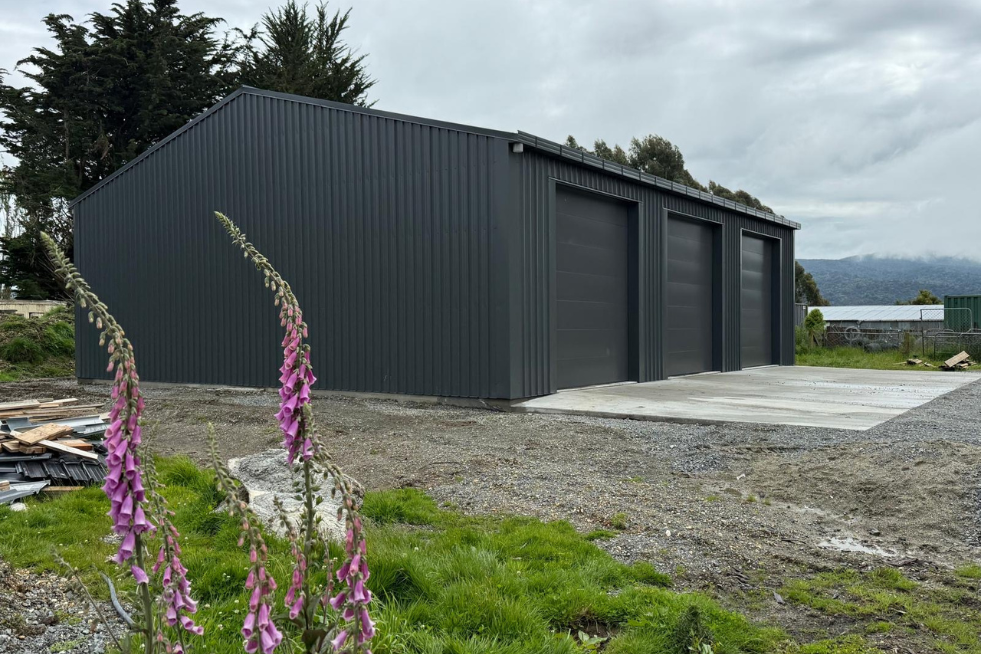 Exterior view of a steel kitset shed in New Zealand with three roller doors, Colorsteel cladding, and concrete slab foundation in a rural setting.